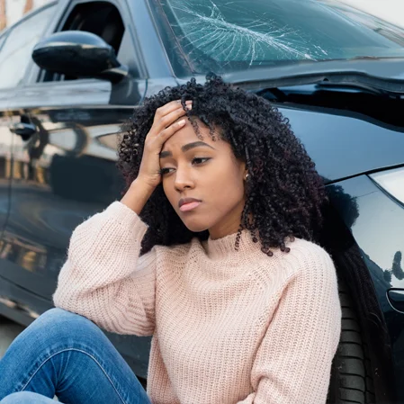 A young woman holding her head while leaning against her damaged car